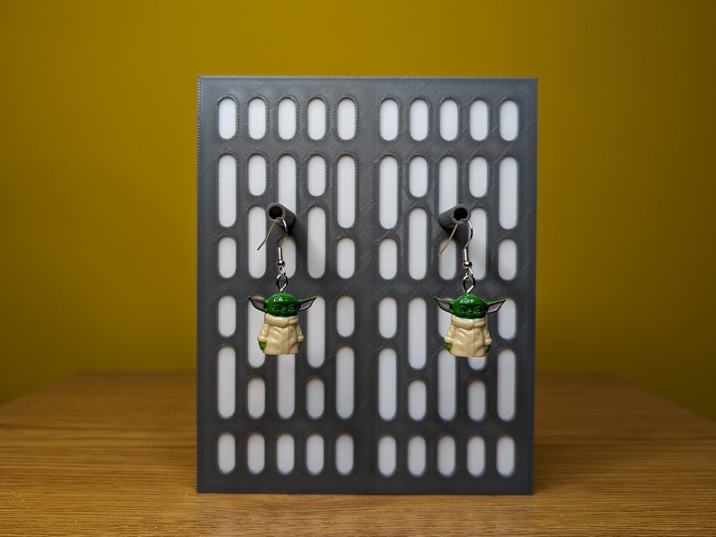 May include: A pair of dangle earrings featuring a green and beige character with large ears. The earrings are hanging from a gray display stand with a white background.