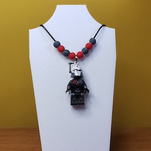 Pode incluir: Um colar preto e vermelho com um pingente de Lego Star Wars de um soldado clone. O colar é feito de cordão preto e contas vermelhas e cinzas.