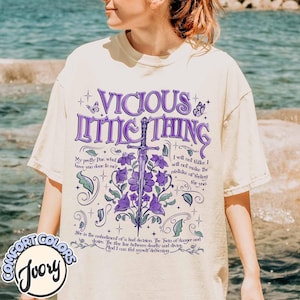 May include: A white t-shirt with a purple and green design featuring the text "Vicious Little Thing" and a quote from the book "The Raven Cycle" by Maggie Stiefvater. The quote reads: "My pretty Poe, what have you done to me? I will not falter. I will not make the mistake of feeling for you. She is the embodiment of a hard decision. The Twin of danger and desire. The fine line between deadly and divine. And I can feel myself drowning."