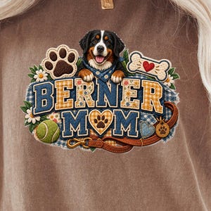 Puede incluir: Una camiseta marrón de manga corta con un estampado gráfico de un perro de montaña de Berna, una huella, un hueso, una pelota de tenis y una correa. El texto "BERNER MOM" aparece en letras azules y amarillas con un corazón sustituyendo la O.