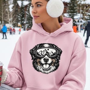 May include: A light pink hoodie featuring a graphic of a dog wearing ski goggles with a mountain scene. The person is wearing white earmuffs and mittens, holding a red plaid coffee cup. The background shows a snowy ski resort.