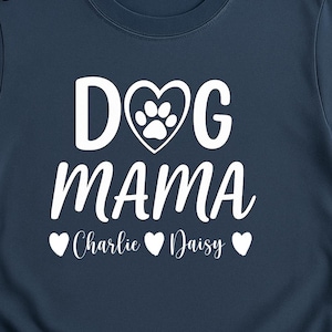 Puede incluir: Camiseta azul marino con las palabras blancas "DOG MAMA", con un corazón con una huella de pata. Debajo, los nombres "Charlie" y "Daisy" con corazones. Gafas de sol y una taza blanca son visibles.