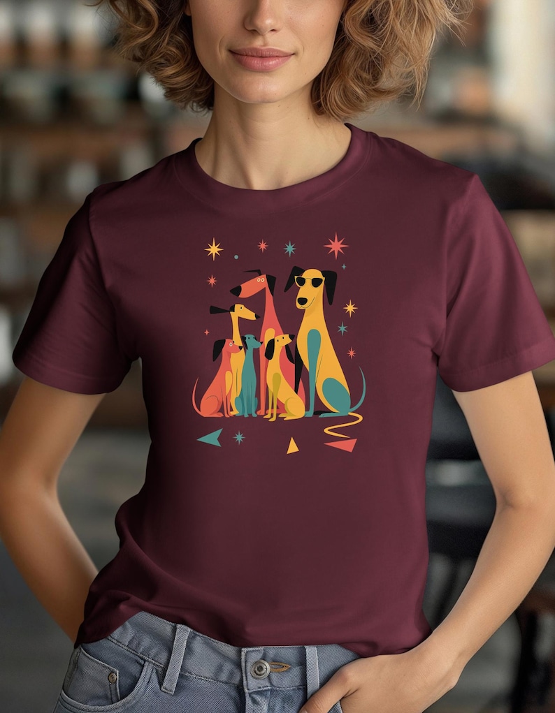 Op de afbeelding: Bordeauxrood T-shirt met een kleurrijke illustratie van gestileerde honden. De honden zijn afgebeeld in geel, blauw en oranje, sommige dragen een zonnebril. Geometrische vormen en sterren omringen de honden.