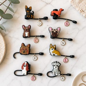 May include: A selection of black cable tidies, each adorned with a different dog breed figurine. The figurines are in various colours, including brown, black, white, and orange. Each tidy has a small, round charm with a letter.