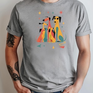Op de afbeelding: Grijs t-shirt met een kleurrijke afbeelding van gestileerde honden. De honden zijn afgebeeld in verschillende kleuren, waaronder geel, rood en blauw, sommige dragen een zonnebril. Geometrische vormen en sterren omringen de honden.