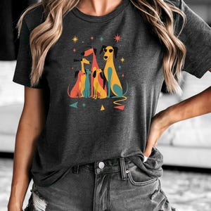 Op de afbeelding: Donkergrijs t-shirt met een kleurrijke afbeelding van gestileerde honden. De honden zijn in verschillende kleuren, waaronder geel, blauw en rood, sommige dragen een zonnebril. Geometrische vormen en sterren omringen de honden.