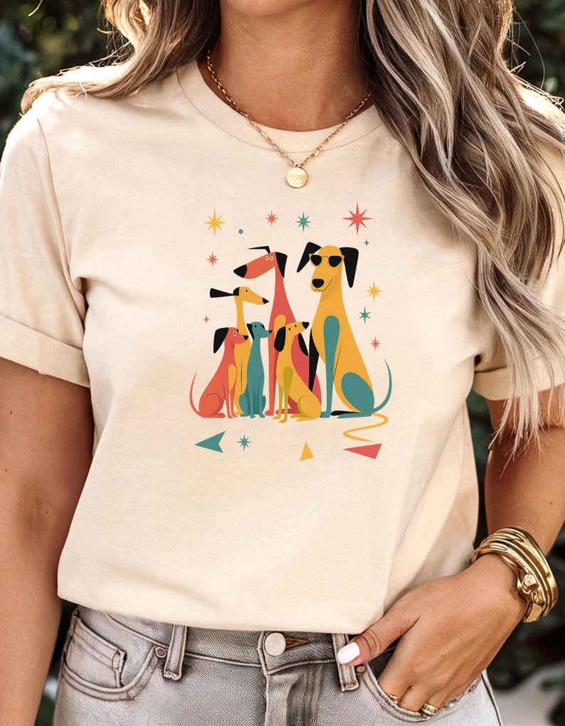 Op de afbeelding: Beige T-shirt met een kleurrijke illustratie van zes gestileerde honden. De honden zijn in tinten geel, oranje, blauw en rood, met ster- en driehoekaccenten. Het shirt heeft korte mouwen.