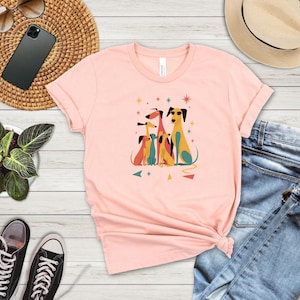 Op de afbeelding: Lichtroze T-shirt met een kleurrijke afbeelding van gestileerde honden. De honden zijn in verschillende kleuren, waaronder geel, rood en blauw, met geometrische vormen en sterren. Het shirt wordt gecombineerd met een blauwe jeans en zwarte sneakers.