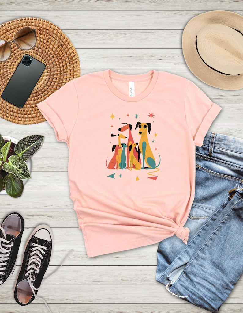 Op de afbeelding: Lichtroze T-shirt met een kleurrijke afbeelding van gestileerde honden. De honden zijn in verschillende kleuren, waaronder geel, rood en blauw, met geometrische vormen en sterren. Het shirt wordt gecombineerd met een blauwe jeans en zwarte sneakers.