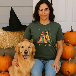 Op de afbeelding: Een vrouw draagt een groen t-shirt met een kleurrijke hondenafbeelding, zittend met een golden retriever en pompoenen. Een zwarte heksenhoed ligt op een hooibaal. De sc&egrave;ne heeft een herfst- of Halloween-thema.