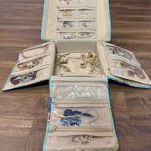 May include: Open jewelry travel case with multiple compartments and clear zippered pockets. The case is light blue with a floral pattern and beige interior. Various pieces of jewelry, including necklaces, earrings, and bracelets, are visible inside.