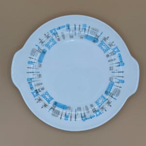 Peut inclure: Un plat de service rond bleu clair avec deux poignées. Le bord présente un motif répétitif d'ustensiles de cuisine stylisés bleus et gris et de formes géométriques. Le plat a un centre bleu clair uni.