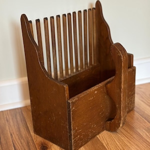 May include: A vintage wooden plate rack with a natural finish. The rack has vertical wooden dowels for plates and a lower compartment. The design includes a handle on the side. The wood shows signs of wear, adding to its antique charm.