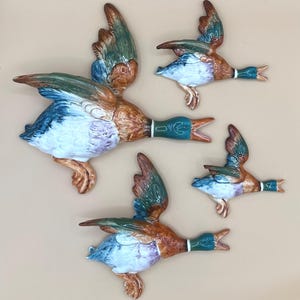 May include: A set of four ceramic wall-mounted mallard ducks in flight, each featuring a glossy finish with painted details in shades of green, brown, blue, and white. The ducks are depicted with open beaks and wings spread in various positions.
