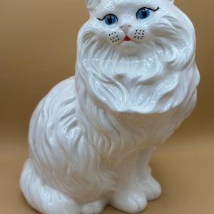 Può includere: Statuetta di gatto in ceramica bianca con occhi blu, baffi neri e bocca rossa. Il gatto ha una pelliccia soffice ed è seduto in posizione eretta. L'oggetto è decorativo.