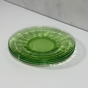 May include: A stack of three round, translucent light green glass saucers. Each saucer features a smooth, flat center surrounded by a wide, raised rim with a repeating radial ribbed pattern that extends to the outer edge of the glass.
