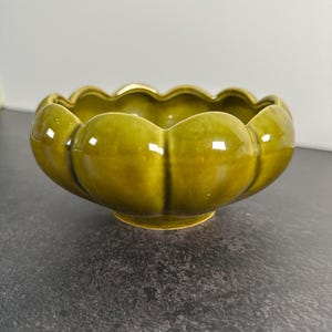 Puede incluir: Un cuenco de cerámica de color verde oliva brillante con un borde festoneado similar a un pétalo y un cuerpo redondeado y bulboso. El cuenco presenta un acabado liso y reflectante y se asienta sobre una pequeña base circular.