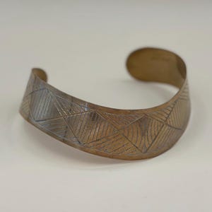 May include: A bronze-colored cuff bracelet with a geometric pattern. The bracelet has a wide band with etched lines forming a series of triangles and parallel lines. The cuff curves slightly, with rounded ends.