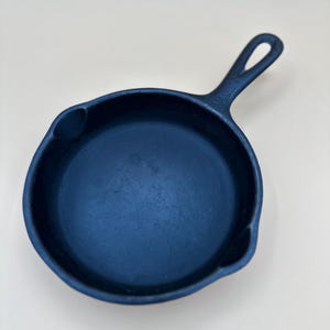 May include: A small, dark blue cast iron skillet with a handle. The skillet is round with a slightly curved rim and a loop at the end of the handle for hanging. The interior of the pan is a darker shade of blue.