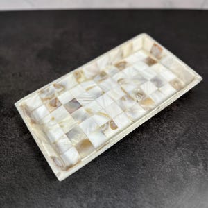 May include: A rectangular tray featuring a mosaic pattern of small, square, iridescent mother-of-pearl tiles. The tiles display varying shades of white, cream, and soft brown, creating a shimmering, reflective surface with a smooth finish.