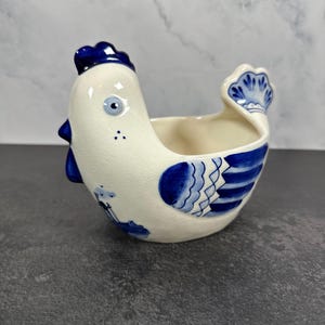 May include: A ceramic chicken-shaped bowl featuring a cream-colored base with hand-painted blue details. The design includes a blue comb, a blue tail, and a small blue windmill illustration on the side. The bowl has a smooth, glossy finish.