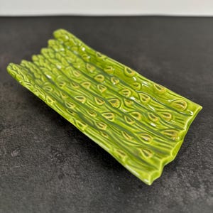 May include: A rectangular ceramic tray featuring a molded design of multiple green asparagus stalks laid side by side. The surface has a glossy finish with raised textures mimicking the natural appearance of asparagus tips and stems in shades of lime.