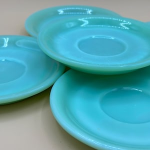 May include: A collection of four turquoise-colored saucers. Each saucer has a circular indentation in the center and a slightly raised rim. The saucers are stacked and arranged, showcasing their smooth, glossy surface.