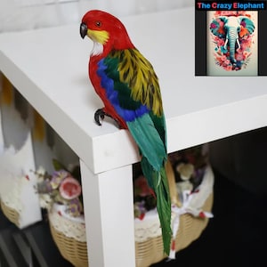 Può includere: Una statuetta di pappagallo realistica e colorata con piume rosse, gialle, blu e verdi. Il pappagallo è appollaiato su un tavolo bianco con un cesto di fiori sullo sfondo.