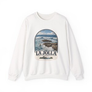 May include: A white crewneck sweatshirt featuring a graphic print of a coastal scene with ocean waves, dark rocks, and a seal in the water. The text LA JOLLA CALIFORNIA is printed below the scene, accompanied by small illustrations of plants.