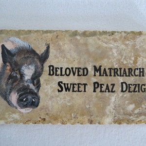 May include: A painted stone plaque with a pig's head on the left side and the text "BELOVED MATRIARCH OF SWEET PEAZ DEZIGNS" on the right side.