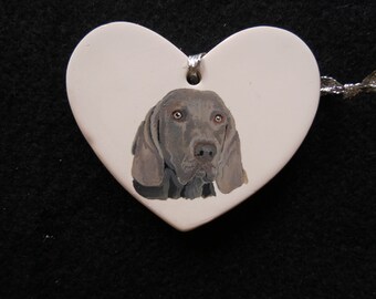 May include: A white ceramic heart-shaped ornament with a painted image of a Weimaraner dog. The dog has gray fur and brown eyes. The ornament has a silver ribbon attached to the top.