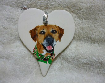 May include: A white ceramic heart-shaped ornament with a painted image of a brown and tan dog wearing a green and red holiday scarf.