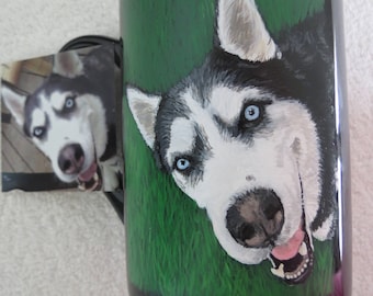 May include: A black ceramic mug with a hand-painted portrait of a Siberian Husky dog. The dog is looking directly at the viewer with its tongue sticking out. The background is a green grassy field.