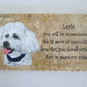 May include: A white dog with a blue collar is painted on a light brown stone. The stone has a message that reads "Leyla, you will be remembered for the 13 years of unconditional love that you shared with us Rest in peace my angel".