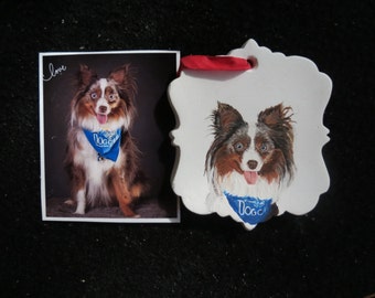 May include: A white ceramic ornament with a painted image of a blue merle Australian Shepherd wearing a blue bandana with the word "Dog" on it. The ornament has a red ribbon attached to the top.