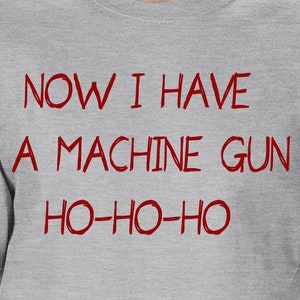May include: Gray t-shirt with the text "NOW I HAVE A MACHINE GUN HO-HO-HO" printed in red.
