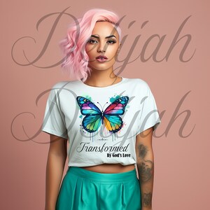 以下が含まれることがあります： カラフルな水彩の蝶のグラフィックと「Transformed By God's Love」のテキストがプリントされた白いTシャツ。