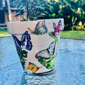 May include: A white ceramic flower pot decorated with colorful butterfly and leaf decals. The pot contains a small green plant. The butterflies are various colors, including blue, pink, and yellow. The pot is sitting on a glass table.
