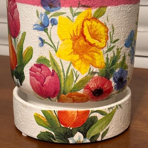May include: White ceramic flower pot with a floral design featuring yellow daffodils, red tulips, and blue flowers. The rim is painted pink, and the pot sits on a matching saucer. A decorative item for home or garden.