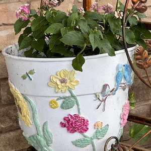 Peut inclure: Un pot de fleurs en céramique blanche orné de reliefs colorés représentant des tournesols jaunes, des pivoines roses, une petite libellule verte et deux oiseaux bleus perchés sur une branche. Il contient une plante verte à fleurs roses.