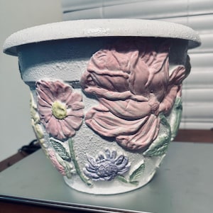 May include: A decorative flower pot with a textured, light gray exterior. The pot features embossed floral designs in shades of pink, purple, and green. The rim of the pot is a lighter shade of gray.