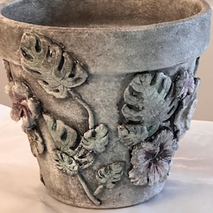May include: A gray, textured ceramic flower pot with embossed tropical leaves and flowers. The pot has a wide rim and a slightly weathered appearance, suitable for indoor or outdoor use. The leaves and flowers are painted in shades of green and purple.