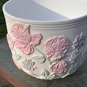 May include: White ceramic planter with embossed pink and purple flowers. The planter has a textured surface and a wide, round opening.