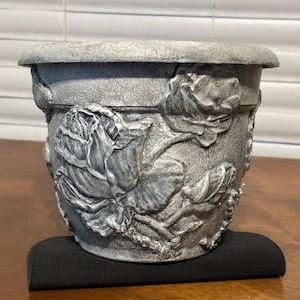 May include: A gray ceramic planter with a raised design of roses and leaves. The planter has a textured surface and a slightly rounded shape.