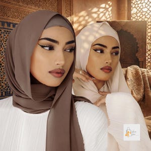 May include: Two hijabs in solid mocha brown and cream colors, worn over white pleated tops. The mocha brown hijab is draped around the head and neck, while the cream hijab is styled in a similar fashion. Both fabrics appear smooth and opaque.