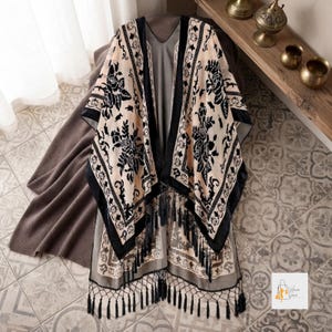 May include: A beige open-front kimono featuring a black velvet floral pattern, a wide black border, and a long fringe hem. The garment has a loose, flowing silhouette with intricate black botanical designs and a solid beige base fabric.