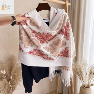 May include: A white scarf with a paisley pattern in shades of brown, red, and green. The scarf has a fringed edge and is draped over a black sweater. The scarf is displayed on a wooden hanger.