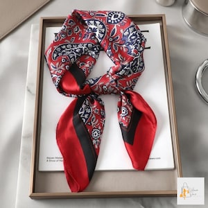 May include: A red and navy blue silk scarf with a paisley pattern. The scarf has a red border with a black stripe. The scarf is folded and draped over a white surface.