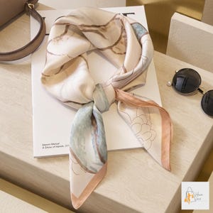 May include: A silk scarf with a floral design in shades of beige, light blue, and peach. The scarf is knotted and lies on a white surface. The scarf has a delicate, feminine aesthetic.