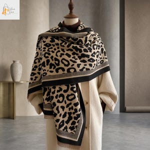 May include: A beige and black leopard print scarf draped over a mannequin. The scarf has a black and gray border and is paired with a cream-colored coat. The scarf's pattern is a classic animal print.